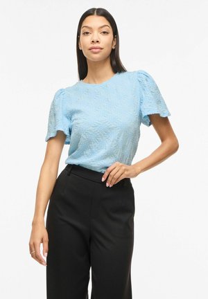 Frau, die eine hellblaue, strukturierte Kurzarmbluse und hoch taillierte schwarze Hose trägt, steht mit einer Hand in der Hüfte.