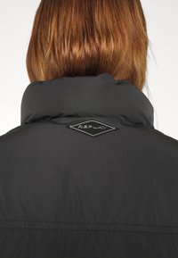 Μαύρο επενδυμένο μπουφάν με ψηλό λαιμό, που διαθέτει ένα λογότυπο σε σχήμα διαμαντιού "REPLAY" στο πίσω μέρος. Λείο υλικό και καθαρές γραμμές.