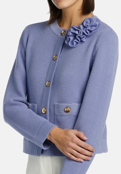 Cárdigan de color púrpura claro con tejido texturizado, cuello redondo, bolsillos frontales y acento floral en el hombro. Presenta botones dorados.