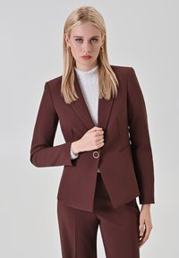 Burgunderfarbener Blazer mit strukturiertem Design, einem einzigen Knopf in Goldfarbe und dezenten Taschen, kombiniert mit einem strukturierten weißen Oberteil darunter.