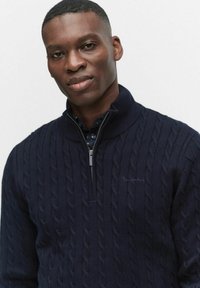 Marinblå stickad tröja med halv dragkedja i kragen, som har en texturerad kabeldesign och en diskret broderad logotyp på bröstet.