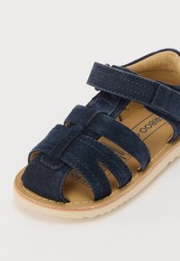 Sandalia de ante azul marino con punta cerrada, múltiples correas y cierre de Velcro. Plantilla acolchada color beige claro y suela cremosa.