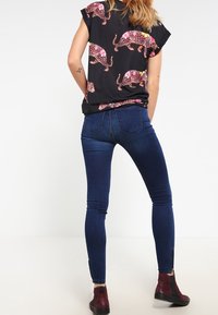 Top negro con flores en patrones rosa y amarillo, combinado con jeans ajustados azul oscuro y botas tobilleras burdeos, visto desde atrás.