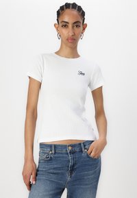T-shirt bianco a righe con maniche corte e scollo rotondo, decorato con il logo ricamato "Guess" in blu, abbinato a jeans di denim blu.