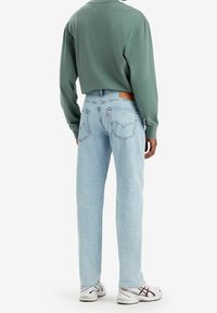 Světlé modré džíny rovného střihu s hnědou koženou nášivkou na zadní straně a jemným opráním, doplněné zelenou košilí s dlouhým rukávem a bílými teniskami.