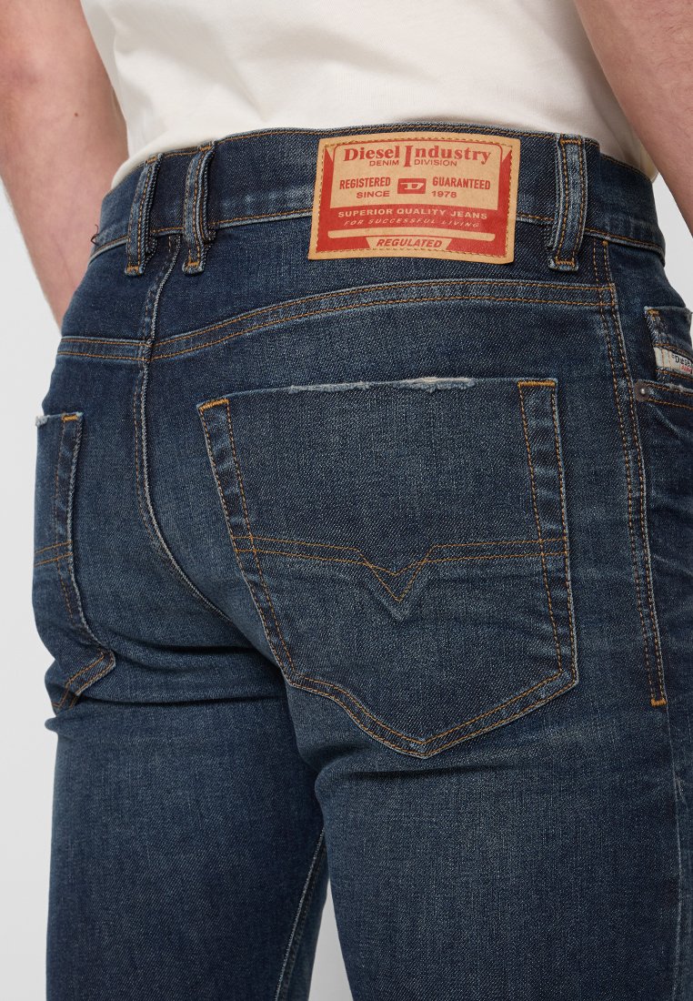 Mann trägt dunkelblaue Diesel-Jeans mit sichtbaren Gesäßtaschen, orangefarbenen Nähten und einem roten Markenlabel am Bund.