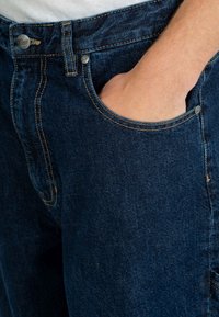 Jeans de mezclilla en azul oscuro con una textura suave. Presenta un bolsillo frontal y un botón de metal, contrastado por costuras más claras alrededor de las costuras.