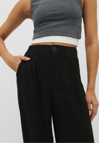 High-waisted black trousers with a button closure, pleats at the front, and side pockets. Smooth, textured fabric with a tailored fit.