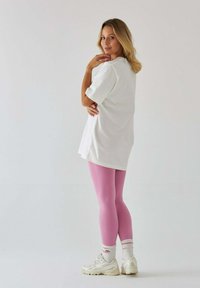 Biała, oversize'owa koszulka, różowe legginsy, białe trampki z akcentami. Modelka stoi bokiem na jasnym tle z delikatną teksturą włosów.