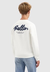Jong persoon met kort blond haar, gekleed in een witte sweatshirt met navy tekst "Ballin" en "Echoes of the Urban Wild" op de achterkant.