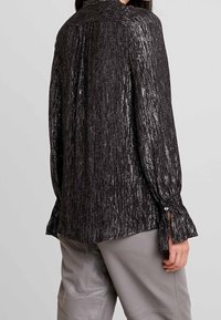 Blusa nera con strisce verticali metalliche, maniche lunghe con lacci ai polsini e un colletto semplice. Dettaglio in tessuto strutturato e lucido.