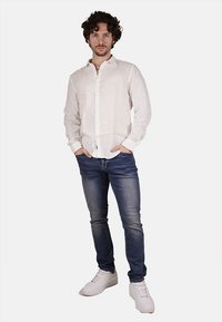Camicia bianca in lino con bottoni, jeans blu e sneakers bianche. La camicia presenta un colletto classico e una trama sottile. Silhouette complessiva aderente.