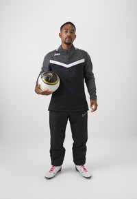 Sportbekleidung in Schwarz und Grau, bestehend aus einem Halbzip-Oberteil mit weißen Akzenten und passenden Hosen. Hält einen Fußball mit goldenen Details.