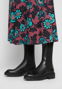 Musta nahkasaapas nilkkamittaisilla varrella, joissa on elastiset sivupaneelit ja teksturoidut kumipohjat. Yhdistetty kukkakuosiset hameet, joissa on turkoosi ja vaaleanpunainen sävy.