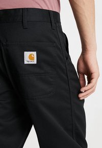 Czarne spodnie z tylną kieszenią z białą metką i pomarańczowym logo; solidna tkanina o gładkiej fakturze i dopasowanym kroju.