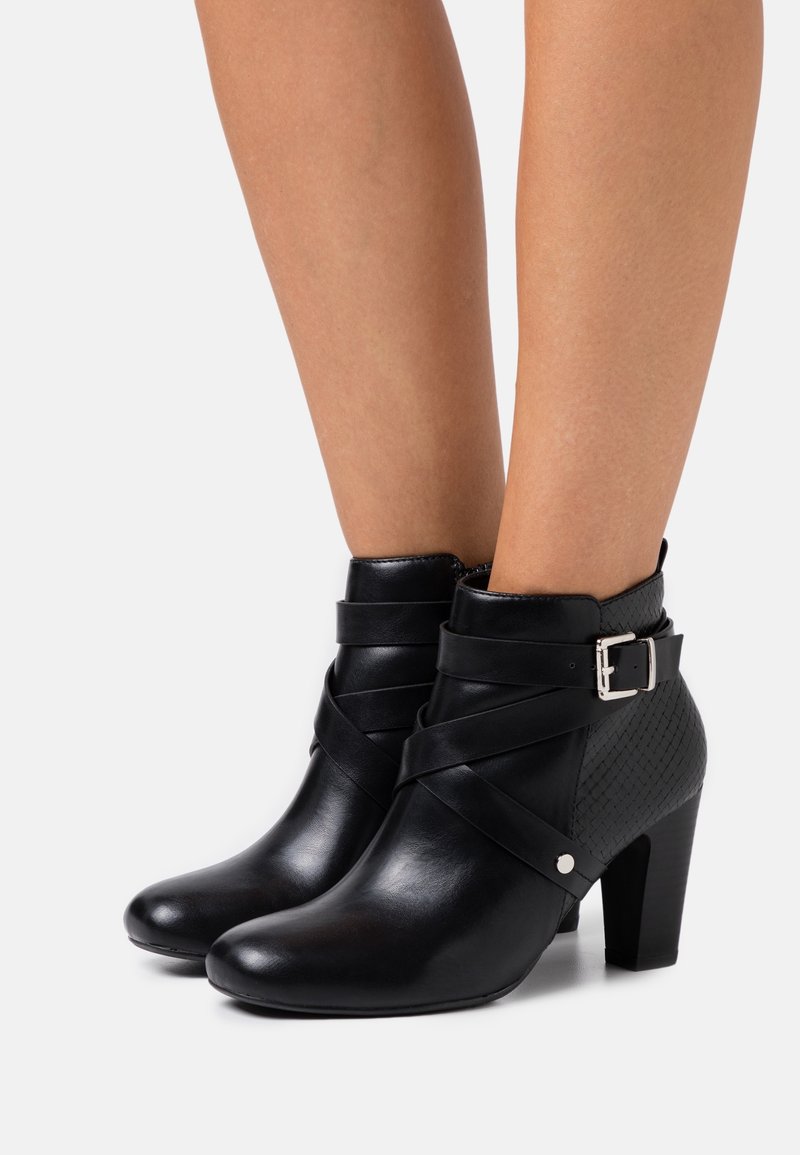 Bottes chevilles en cuir noir avec des sangles croisées, détail de boucle argentée, contrefort texturé et talons carrés portées sur jambes nues.