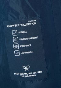Etiqueta de tela azul marino de la Colección de Ropa Exterior de Silbon. Presenta iconos y texto que indican durabilidad, comodidad, resistencia al viento y ligereza.