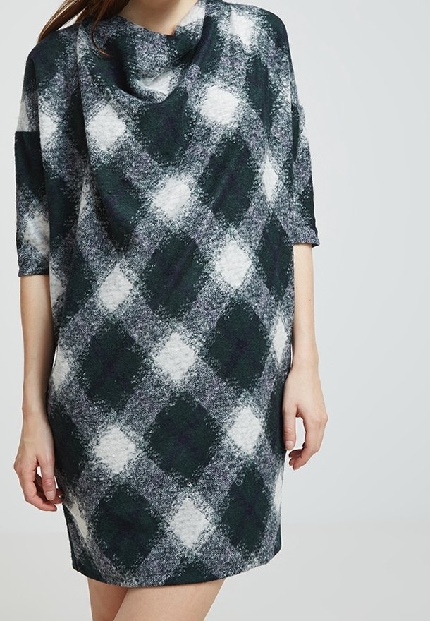 Femme portant une robe ample mi-longue à manches trois-quarts, arborant un motif à carreaux losanges en noir, blanc et gris.