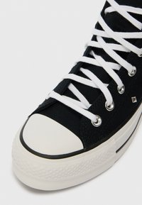 Suurendatud vaade mustale kõrgele kanvas tennisekingale, millel on valge kummine ninaosa ja valged paelad valgel taustal.