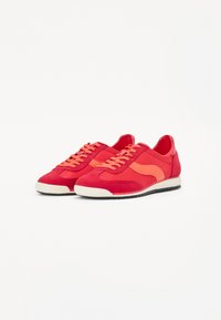 Red canvas sneakers with tonal suede accents, featuring a white rubber sole, round toe, and flat laces. Distinctive side design detail.