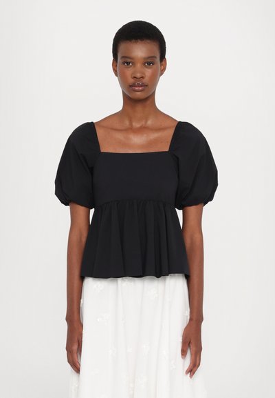 Haut noir à manches bouffantes avec un décolleté carré et des détails plissés à la taille, fait en tissu léger, porté sur une jupe blanche brodée.