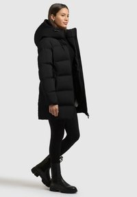 Cappotto imbottito nero con colletto alto, caratterizzato da una chiusura con zip, fodera interna e vestibilità rilassata; indossato con leggings neri e stivaletti neri.