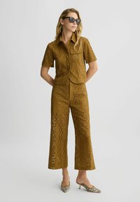Braunes Leinenhemd mit Lochstickerei und passenden weit geschnittenen Hosen, die kreisförmige Ausschnitte aufweisen. Glänzende Sonnenbrille und gemusterte High-Heels runden das Outfit ab.