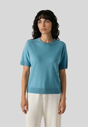 Vrouw met donker krullend haar, die een blauw gebreid topje met korte mouwen en een witte broek draagt tegen een effen lichtgrijze achtergrond.