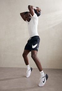 Mustat urheilushortsit, joissa on valkoinen Nike-logo, yhdistettynä valkoiseen tiukkaan paitaan ja valko-mustiin kenkiin. Henkilö pitää painopalloa pään yläpuolella.