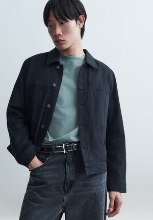 Νεαρό άτομο με μαύρο σακάκι με κουμπιά πάνω από πράσινο πουκάμισο, μαύρη ζώνη με καρφιά και σκούρο τζιν, με το χέρι στην τσέπη, κοιτάζοντας πλάγια.