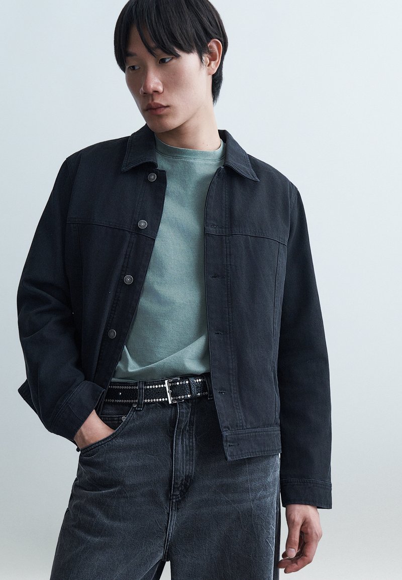 Νεαρό άτομο με μαύρο σακάκι με κουμπιά πάνω από πράσινο πουκάμισο, μαύρη ζώνη με καρφιά και σκούρο τζιν, με το χέρι στην τσέπη, κοιτάζοντας πλάγια.