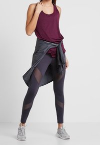 Bordowy top na ramiączkach i ciemne legginsy z siateczkowymi akcentami. Szara lekka kurtka związana w talii. Szare buty sportowe z teksturowaną podeszwą.