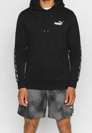 Mężczyzna ubrany w czarną bluzę Puma z kapturem, z logo na piersi i taśmą z nadrukiem na rękawach, w połączeniu z szarymi, wzorzystymi szortami Puma.