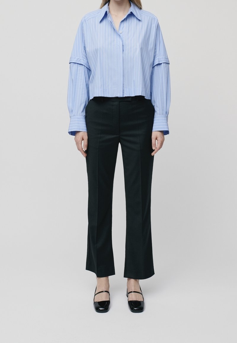 Chemise bleue à rayures recadrée avec des manches oversize, associée à un pantalon noir sur mesure et des chaussures à talons blocs noires. Design simple et structuré.