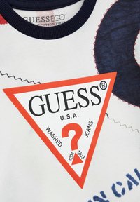 T-shirt bianco in cotone con un grande logo triangolare "GUESS" rosso e nero. Elementi notevoli includono un punto interrogativo e dettagli di cucitura.