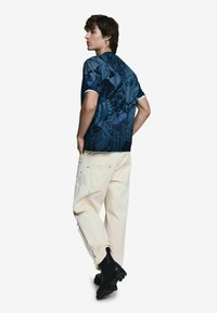 Modré vzorované tričko s krátkym rukávom a bielym golierom, skombinované s krémovými nohavicami; modelka obutá v čiernych topánkach, zobrazená zozadu.