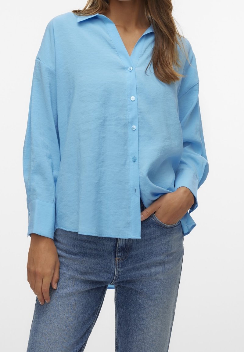 Femme portant une chemise ample bleu clair à boutons avec manches longues et un jean bleu, une main dans la poche et l'autre détendue sur le côté.