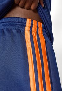 Tuvumā tumši zilas sporta bikses ar trim spilgti oranžām svītrām un elastīgu jostu, roka paceļot tumši zilu krekli, lai atklātu vidukli.