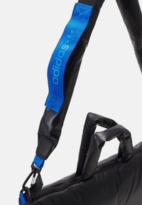 Sac d'épaule noir à finition brillante, avec une sangle bleue à logo et des poignées rembourrées. Comprend une fermeture éclair et des coutures contrastées.