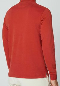 Uomo che indossa una camicia a maniche lunghe color ruggine, visto di schiena, abbinata a pantaloni chiari su uno sfondo semplice.