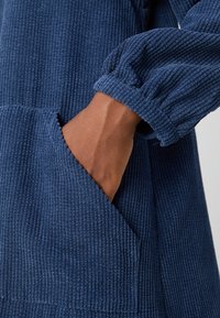 Main insérée dans la poche avant d'un vêtement à manches longues en velours côtelé bleu avec un détail de poignet élastique froncé.