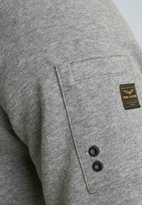 Grauer Pullover mit strukturierter Oberfläche, ausgestattet mit einer Tasche mit einem schwarzen Etikett und zwei schwarzen Druckknöpfen am Ärmel.