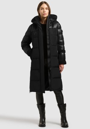 Mujer de pie con un abrigo largo acolchado negro con capucha, pantalones negros y botas negras hasta la mitad de la pantorrilla, sobre un fondo gris claro liso.