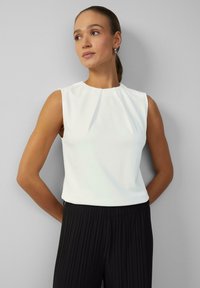 White, sleeveless blouse with a round neckline and pleated detailing, paired with black pleated trousers. Smooth fabric texture.