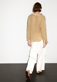 Maglietta a maniche lunghe color marrone chiaro, a coste, con colletto, abbinata a pantaloni bianchi larghi e sandali con accenti rossi, vista da dietro.