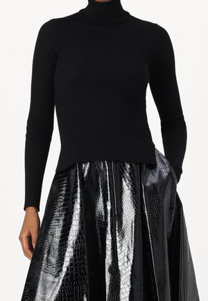 Žena nosící černý žebrovaný rolák a lesklou černou koženou sukni s texturou, s jednou rukou v kapse.