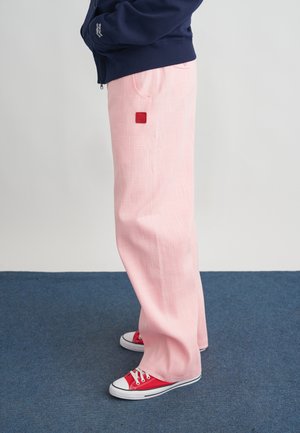 Persona con pantalones sueltos de color rosa con un parche cuadrado rojo, zapatillas rojas y blancas, y chaqueta azul marino, de pie sobre una alfombra azul.