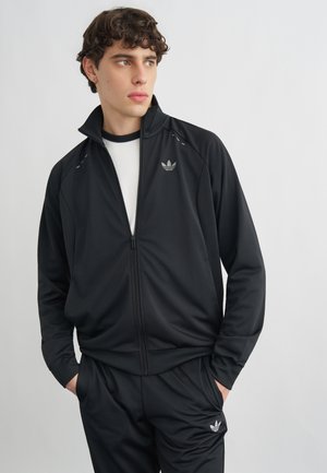 Giovane uomo con capelli ricci che indossa una giacca Adidas nera con zip e pantaloni abbinati, mani nelle tasche, che guarda di lato contro uno sfondo semplice.