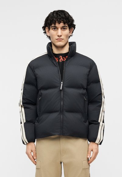 Ung mand med krøllet mørkt hår iført en sort dunjakke med hvide striber på ærmerne og beige cargobukser, stående og vendt fremad.