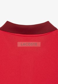 Röd polo-shirt med en mörkröd ribbad krage och en subtil texturerad yta. Har en 'LACOSTE'-logotyp sydd på tyget.